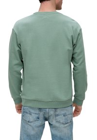 Sudadera de algodón verde con cuello y puños de canalé, que presenta una textura suave. Combinada con unos vaqueros de mezclilla azul desgastados.