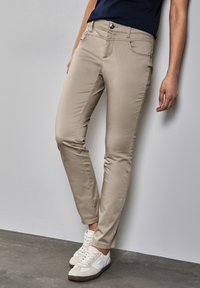 Valkoiset, slim-fit housut viidellä taskulla, nappikiinnitys ja huomaamaton ompelu, yhdistettynä valkoisiin tennareihin harmaalla lattialla.