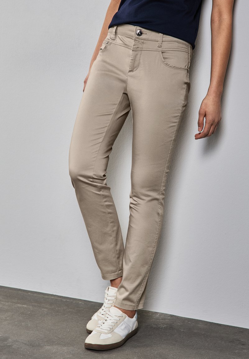 Valkoiset, slim-fit housut viidellä taskulla, nappikiinnitys ja huomaamaton ompelu, yhdistettynä valkoisiin tennareihin harmaalla lattialla.