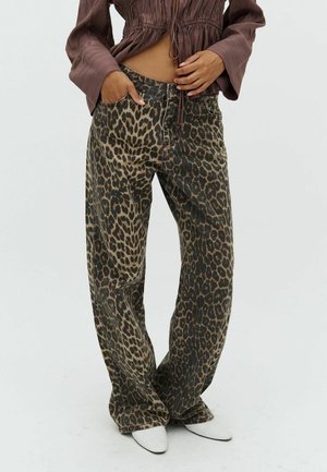 Woman wearing brown ruched top and loose-fitting leopard print pants with white pointed shoes, standing with one hand in pocket.