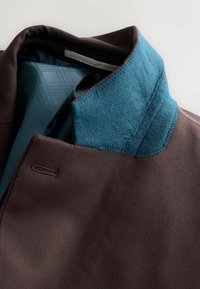 Bruine buitenstof jasje met een gestructureerde, teal wolkraag en een blauw patroon voering. Heeft een gesloten voorkant met een enkele stikdetail.