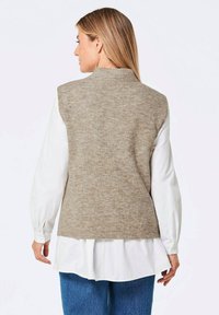 Gilet en tricot marron clair avec un col haut, porté sur un chemisier blanc. Présente une texture côtelée et des variations de couleur subtiles.