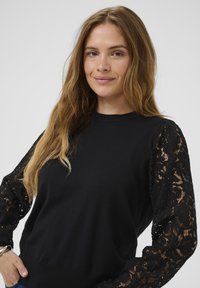 Sieviete ar gariem gaiši brūniem matiem, ģērbusies melnā blūzē ar mežģīņu ziedu rakstu piedurknēm, nedaudz smaidot pret vienkāršu gaišu fonu.