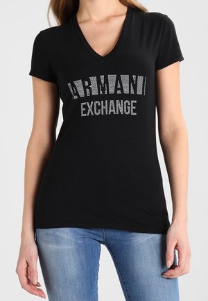 Mujer vistiendo una camiseta negra de corte en V de Armani Exchange con un logo de strass, combinada con jeans de mezclilla azul claro, frente a un fondo liso.