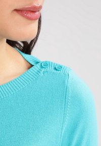 Light blue sweater featuring a ribbed neckline and two small buttons near the shoulder. Made from a soft, knit material.