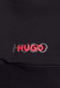 Svart sweatshirt-tyg med ribbad textur. Har en flerfärgad "HUGO"-logotyp i röd och grå, som är placerad framträdande på framsidan.
