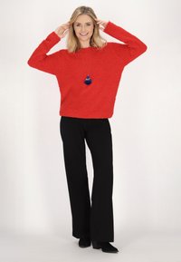 Roter, flauschiger Strickpulli mit weitem Schnitt, kombiniert mit schwarzen, weit geschnittenen Hosen und Absatzstiefeln. Eine blaue Quasten-Halskette sorgt für Details.