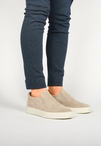 Béžové slip-on boty vyrobené z surové kůže s texturovaným povrchem a elastickými bočními detaily, doplněné o tmavě modré úzké kalhoty.