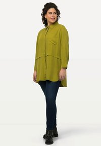 Camisa verde caqui de botón con cuello y dos bolsillos, con un dobladillo curvo y un corte relajado, combinada con jeans oscuros y botas negras hasta el tobillo.