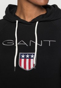 Sudadera negra con cordones blancos, presenta un logo de escudo bordado en rojo, blanco y azul. Tela suave y mangas raglán.