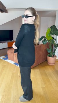 Czarny sweter z długim rękawem o luźnym kroju, w połączeniu z czarnymi spodniami i beżowymi klapkami. W tle znajduje się roślina i kanapa.
