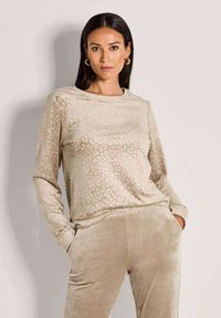 Vrouw met lang donker haar die gouden oorbellen draagt, een beige panterprint lange mouw top en bijpassende beige fluwelen broek met haar handen in de zakken.