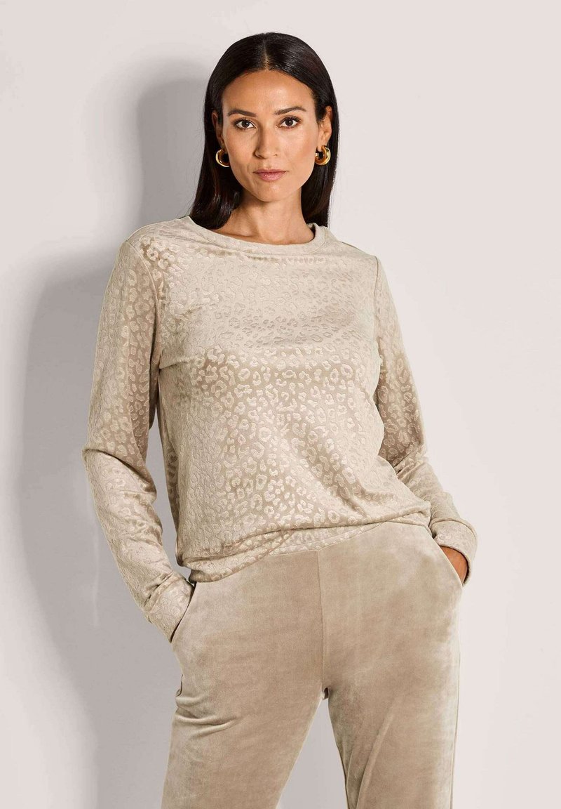 Vrouw met lang donker haar die gouden oorbellen draagt, een beige panterprint lange mouw top en bijpassende beige fluwelen broek met haar handen in de zakken.