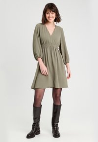 Robe verte au-dessus du genou avec un décolleté en V et une taille élastique. Avec des manches bouffantes longues et un tissu texturé. Associée à des bottes noires montant jusqu'au genou.