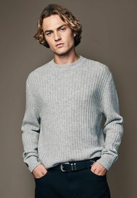 Light gray ribbed knit sweater with long sleeves and a crew neck, worn over dark pants. Features side split hems and a smooth texture.