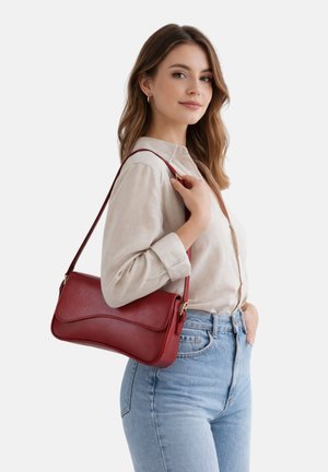 Jonge vrouw in beige blouse en blauwe spijkerbroek met een rode leren schoudertas met een gebogen klep, staand tegen een witte achtergrond.