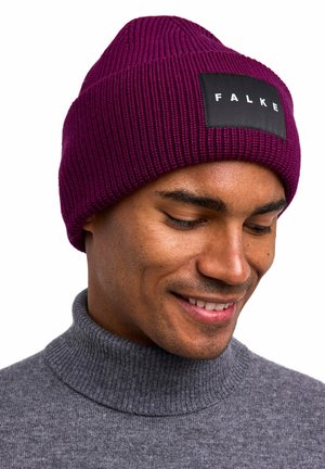 Man smiling, wearing ribbed purple knit beanie with "FALKE" label and gray turtleneck sweater, looking downward against white background.