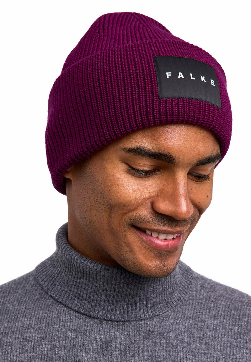 Man die glimlacht, met een geribde paarse gebreide muts met "FALKE" label en een grijze coltrui, kijkend naar beneden tegen een witte achtergrond.