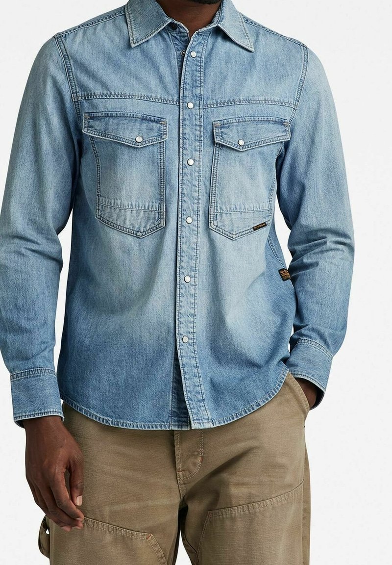 Lichtblauw denimshirt met lange mouwen, drukknoopsluiting en twee borstzakken. Het shirt heeft een kraag en een iets gebogen zoom.
