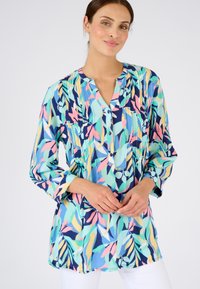 Blusa colorida con un estampado de hojas en azul, verde, rosa y amarillo. Tiene un cierre frontal con botones y mangas de tres cuartos.
