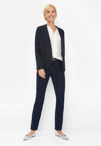 Blazer bleu marine sur une blouse blanche, associé à un pantalon en denim foncé et des ballerines argentées. Textures lisses, coupe ajustée et détails de conception simples.