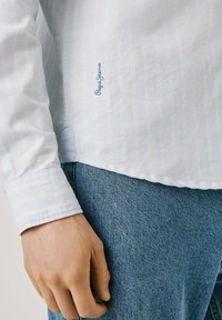 Gaiši zila garas piedurknes krekls ar dzeltenām svītrām un zilu izšuvumu kreisajā pusē, apvienots ar teksturētām zilām džinsu biksēm.