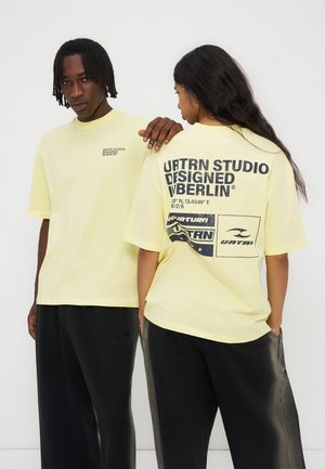 Deux personnes portant des T-shirts oversize jaune clair avec des textes et graphiques noirs, associés à des pantalons noirs amples, debout contre un mur uni.