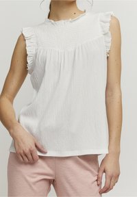 Vit ärmlös topp med volangdetaljer, strukturerad tyg, rundad halsringning och lös passform, kombinerad med ljusrosa byxor.