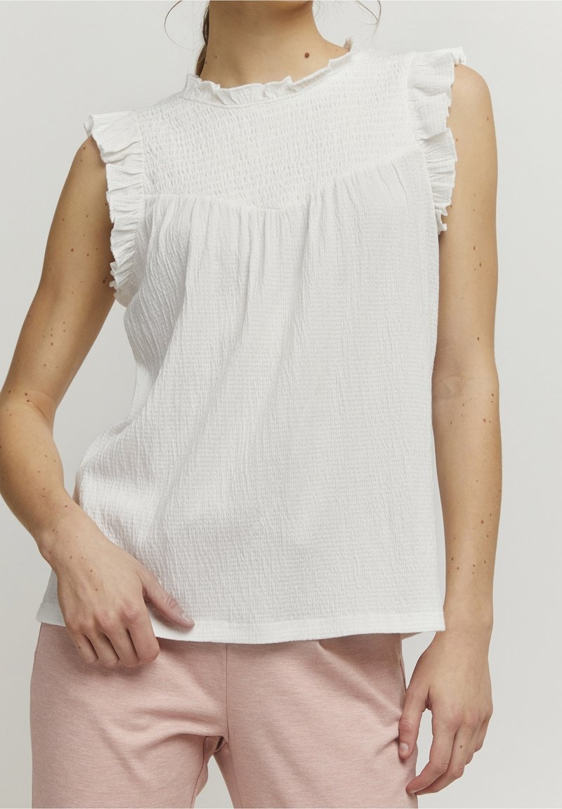 Vit ärmlös topp med volangdetaljer, strukturerad tyg, rundad halsringning och lös passform, kombinerad med ljusrosa byxor.