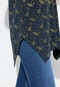 Blusa azul marino con un patrón de frutas amarillas, con un dobladillo en punta y aberturas laterales, combinada con jeans azules. Textura de tela suave visible.