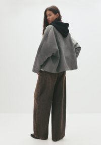 Jachetă oversized gri cu glugă neagră, asociată cu blugi largi de culoare maro închis. Textură de țesătură netedă, design minimalist, fără modele vizibile.