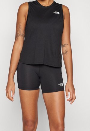 Kobieta ubrana w czarny bezrękawnik i dopasowane obcisłe szorty z logo The North Face, pokazująca tatuaże na obu ramionach.