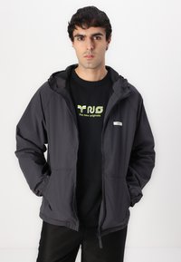 Graue leichte Jacke mit Kapuze, Reißverschluss vorne und elastischen Bündchen, getragen über einem schwarzen T-Shirt mit einem grafischen Druck in Neonfarben.