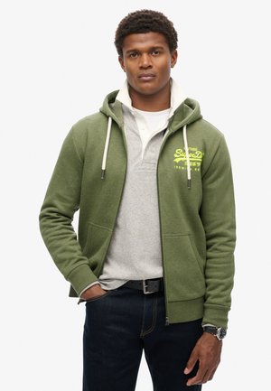 Groene ritsjack met drawstrings, met een geel logo op de borst, gedragen over een grijze polo. Gepaard met donkere jeans en een zwarte riem.