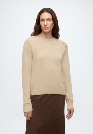 Femme aux longs cheveux ondulés porte un pull beige avec des boutons dorés à l'épaule et une jupe marron foncé sur fond blanc.