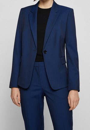 Persona vistiendo un blazer azul marino a medida y pantalones a juego sobre una camiseta negra de cuello redondo, de pie frente a un fondo liso y claro.