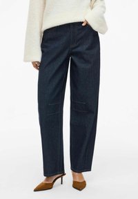 Dark blue denim cargo pants with reinforced pockets, straight leg design, and contrasting yellow stitching, paired with brown heeled mules.