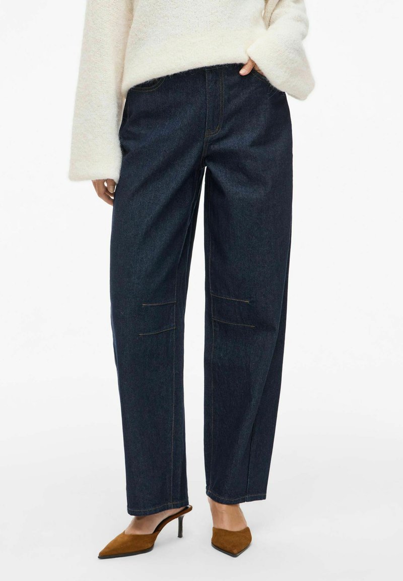 Dark blue denim cargo pants with reinforced pockets, straight leg design, and contrasting yellow stitching, paired with brown heeled mules.