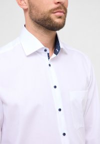 Camicia bianca con bottoni e collo blu navy a pois. Presenta bottoni scuri, una tasca singola sul petto e una trama liscia.