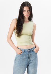 Top ajusté sans manches de couleur vert clair avec un col rond et une texture froncée, associé à un jean bleu déchiré taille haute.