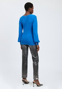 Frau steht mit dem Rücken zu uns und trägt einen leuchtend blauen Pullover, metallisch silberne Hosen und schwarze High-Heels mit offenen Fersen und Verzierungen.