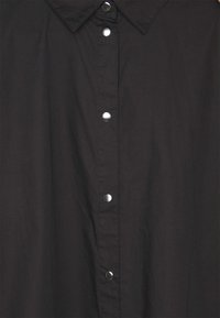 Devant de chemise noire avec col pointu et cinq boutons-pression argentés visibles en ligne verticale.