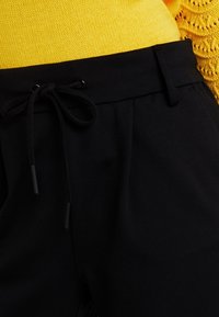 Černé kalhoty s elastickým páskem, které jsou kombinovány se žlutým pleteným topem s texturovaným krajkovým vzorem na rukávech.