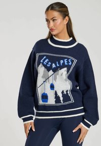 Maglione navy con accenti bianchi e azzurri, che presenta una scena montana, funivie e il testo "LES ALPES" in caratteri in grassetto. Texture morbida.