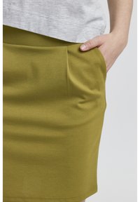 Jupe vert olive en tissu doux, dotée de poches latérales et d'une texture lisse. Assortie avec un haut gris partiellement visible au-dessus.