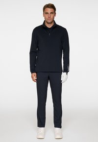 Pullover lungo a maniche nere navy scuro con colletto a mezza zip, abbinato a pantaloni coordinati e sneakers bianche; include un guanto da golf bianco sulla mano sinistra.