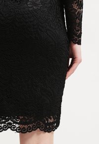 Robe en dentelle noire avec motifs floraux, ourlet festonné et manches en dentelle transparente ; vue de la taille au genou du côté droit d'une personne.