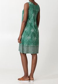 Robe sans manches verte avec un motif paisley, un tissu texturé et un ourlet incurvé. Associée à des sandales à glisser marron sur un fond blanc.