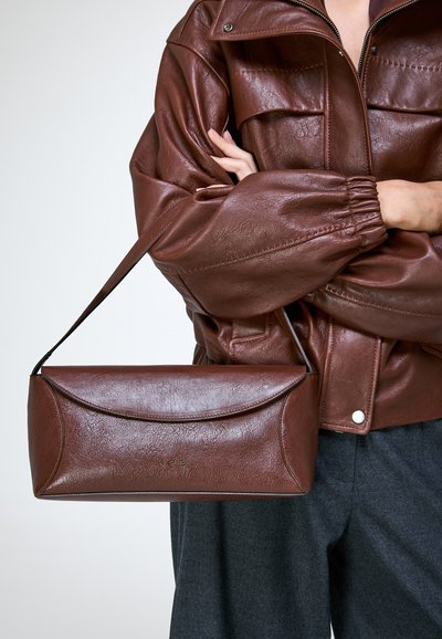 Personne portant une veste en cuir marron tenant un sac à main en cuir marron assorti avec les lettres VSP sur le devant.