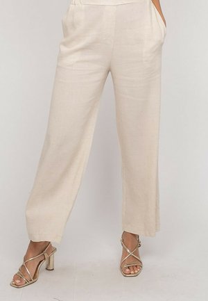 Pantalon beige à jambes larges porté avec des sandales à talons hauts dorées à lanières, les mains placées de manière décontractée dans les poches.